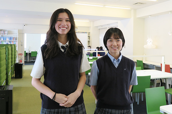 広々と明るい図書室に並び、笑顔を見せる女子生徒2名