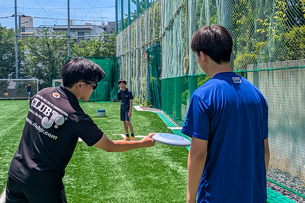 人工芝のグラウンドで、フライングディスク(フリスビー)を手に講師から投げ方の指導を受ける生徒