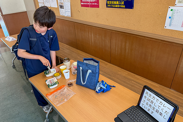 テーブルで手作りカレーを盛り付ける生徒。テーブル上のタブレット画面には「先生方でカレーを試食してくださる先生募集しています」