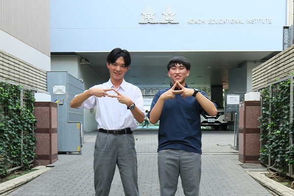 N.S.くん、K.K.くんが校舎を背景に、指で三角を作って立っている様子