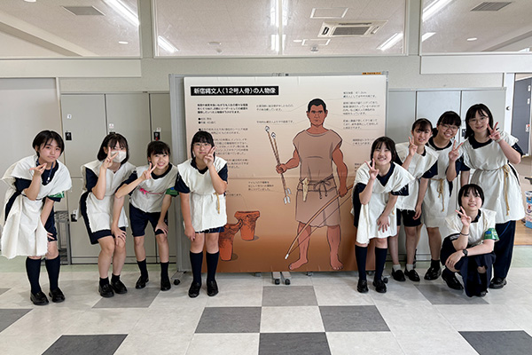 オープンキャンパスにて、新宿縄文人のパネルの前で、パネルと同じ服装をして笑顔を見せる生徒たち。