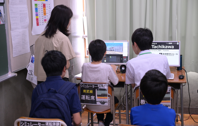「鉄道運転シミュレーター」を操作する小学生と説明している高輪中学高等学校旅行・鉄道研究部部員の写真